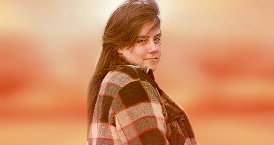 A young woman smiles in a flannel shirt, outdoors amid warm tones.
