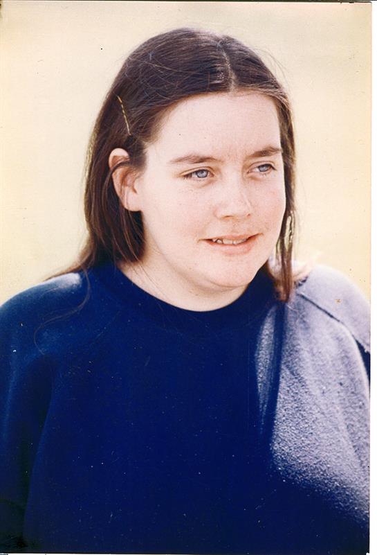 A woman stands outdoors with long hair, wearing a blue sweater, smiling lightly in the sun.