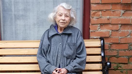 An elderly woman with white hair is seated on a bench, enjoying a peaceful moment outdoors.