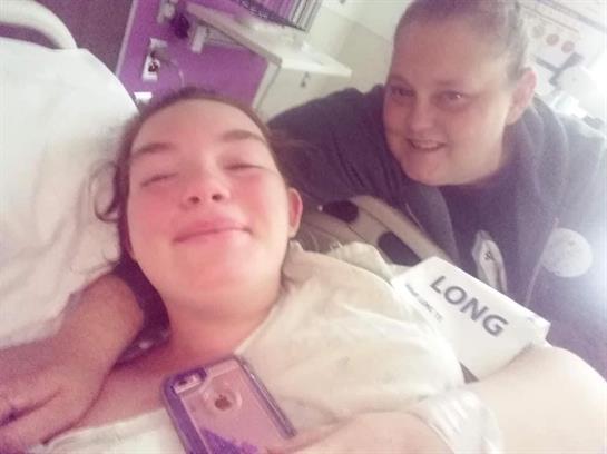 Young woman smiles in a hospital bed, supported by a friend during her recovery.