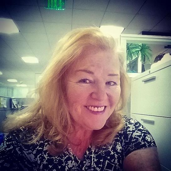 A woman with long blonde hair smiles warmly while seated at her office desk.