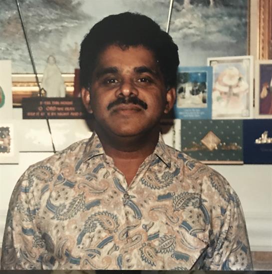 A man with a mustache poses confidently in front of a wall filled with artwork and postcards.