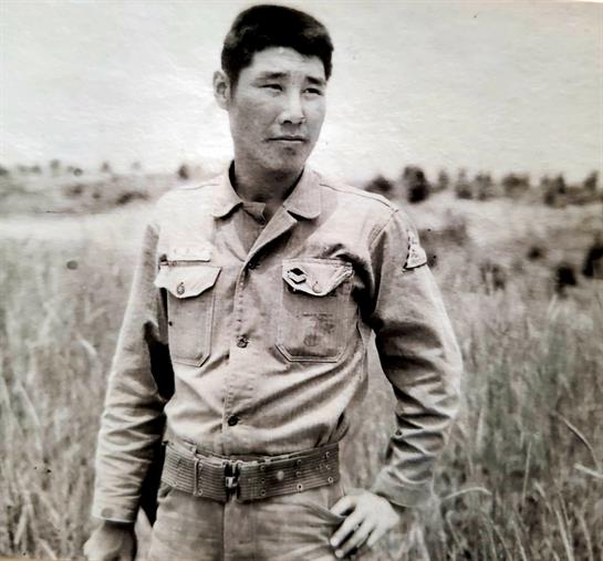 Soldier poses in military uniform, showcasing a serious expression in a field of grass.