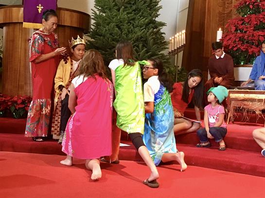 Kids in colorful costumes perform on stage in a church with decorations and a Christmas tree nearby.