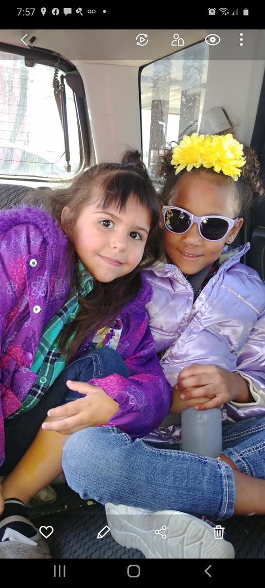 Two girls sit closely together, wearing colorful jackets and sunglasses, smiling happily.