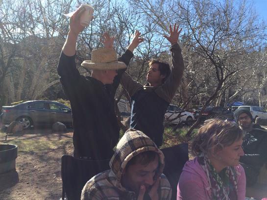 Two men joyfully raise their hands while others gather nearby in a sunny outdoor environment.