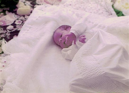 A baby lies wrapped in a white blanket, calmly resting on a plush surface at home.