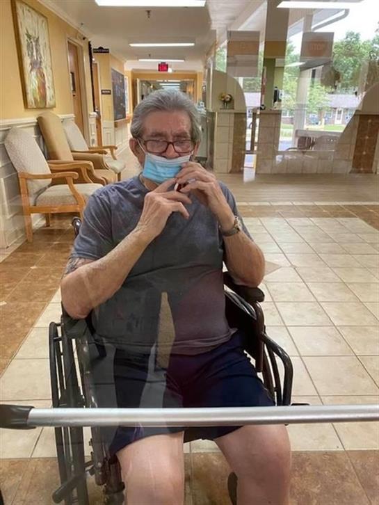 Senior man in a wheelchair is adjusting his mask while sitting indoors in a care facility.