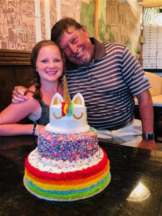 A joyful celebration features a girl and a man smiling proudly with a bright unicorn cake.
