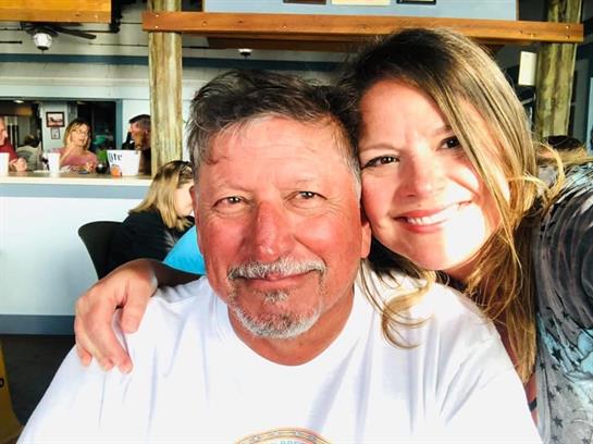 Couple smiles joyfully at a coastal restaurant while friends gather in the background.