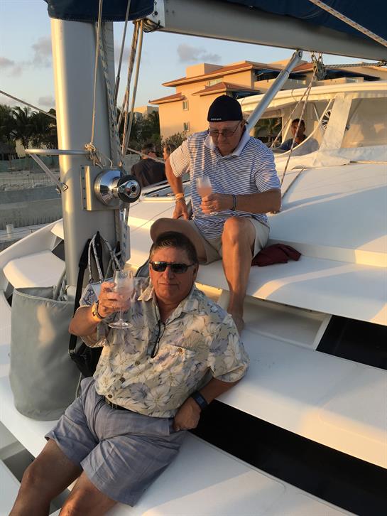 Two friends relax on a yacht, holding drinks and enjoying the sunset over the water.