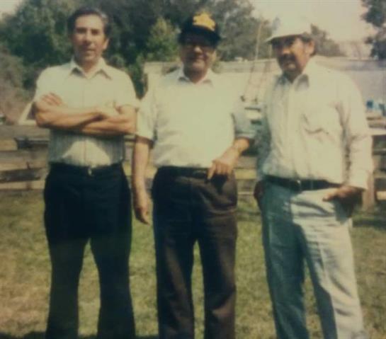 Three men standing side by side in a grassy area, smiling and dressed casually in hats.