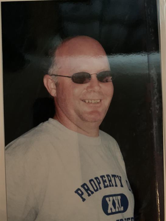 A man with bald head and sunglasses smiles warmly while wearing a casual shirt.