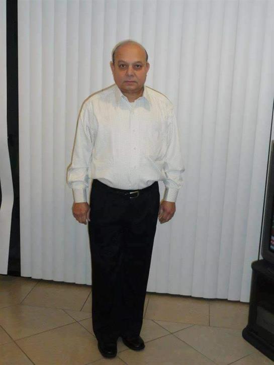 A man stands upright indoors, dressed in a white shirt and black trousers, looking ahead.