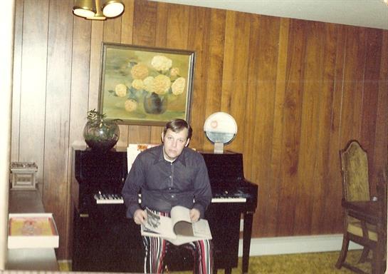A man sits casually reading a magazine next to a piano, surrounded by wooden walls and decor.