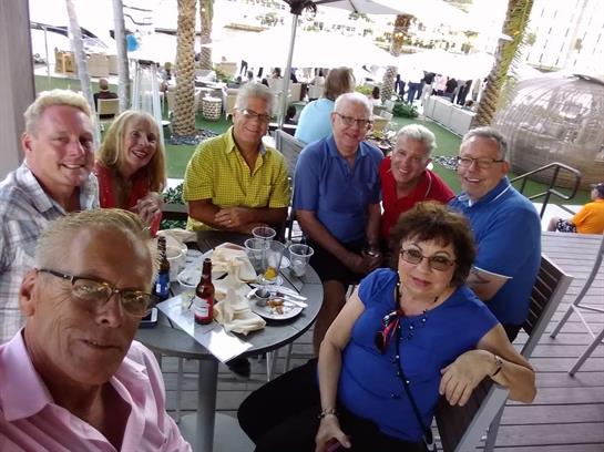 Friends gather around a table sharing laughter, drinks, and food in a cheerful outdoor setting.