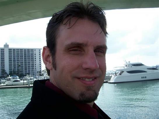 A man stands by the water with boats and buildings visible in the background, smiling happily.