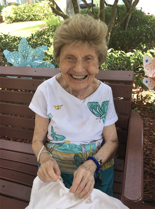 Smiling elderly woman sits on a park bench, enjoying the sunny weather and colorful decorations.