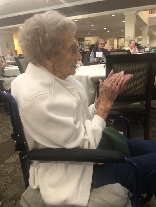 An elderly woman in a wheelchair claps happily as other residents socialize around her.