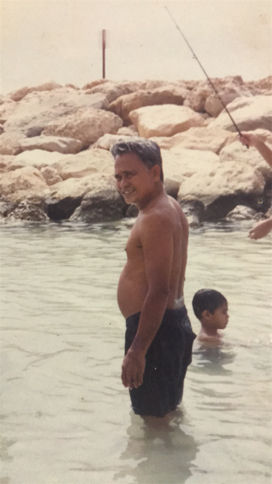 An elderly man smiles in shallow water while a child plays nearby on a warm beach day.