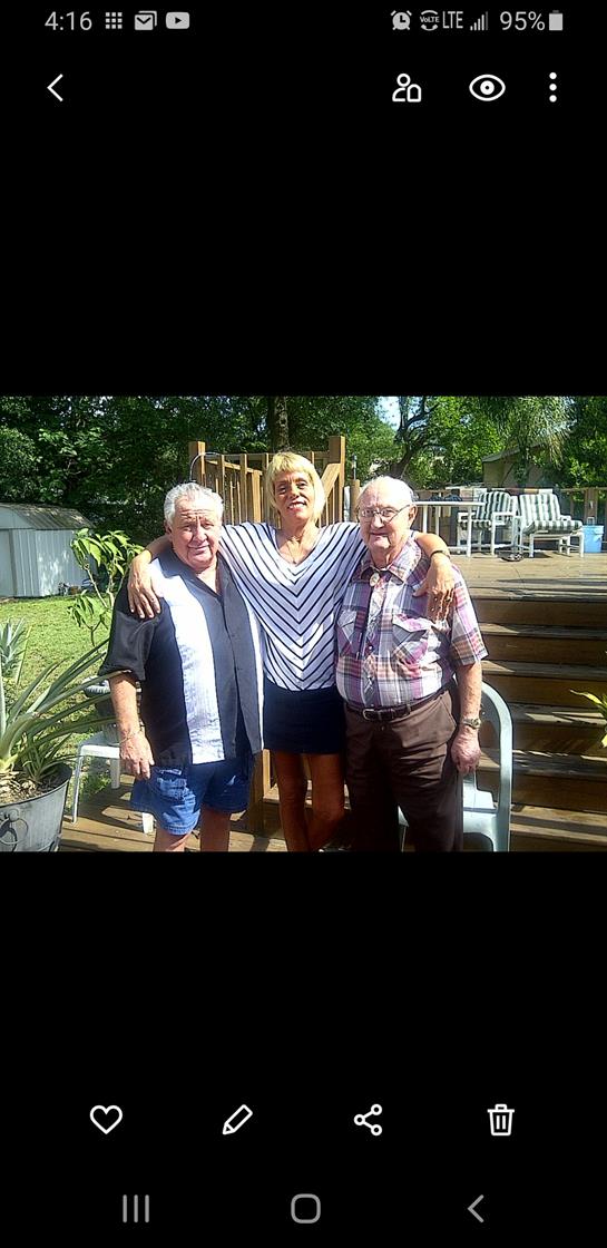 Three friends stand closely together, smiling warmly in an outdoor backyard setting on a sunny day.