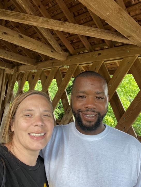 Two friends pose for a cheerful selfie under a rustic wooden structure, smiling happily.