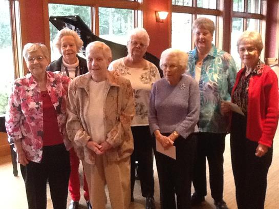 Senior women stand together smiling at a community center event featuring a piano.