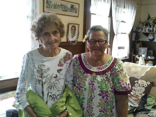 Two smiling elderly women wear vibrant outfits and stand together in a warm, inviting room.