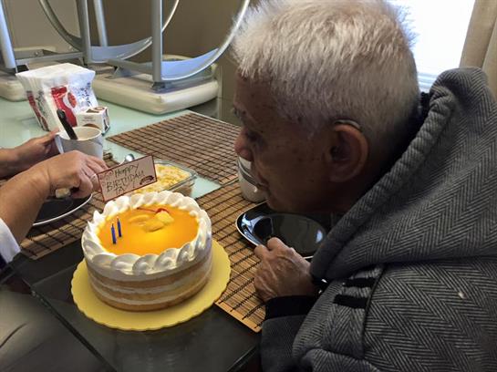 An elderly man leans towards a cake, ready to blow out candles with friends around him.