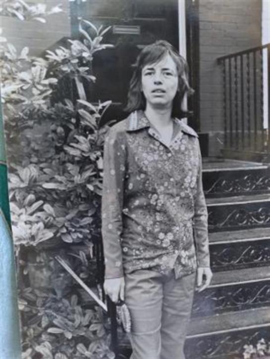 Individual with long hair stands outside a house, dressed in a floral patterned shirt and jeans.