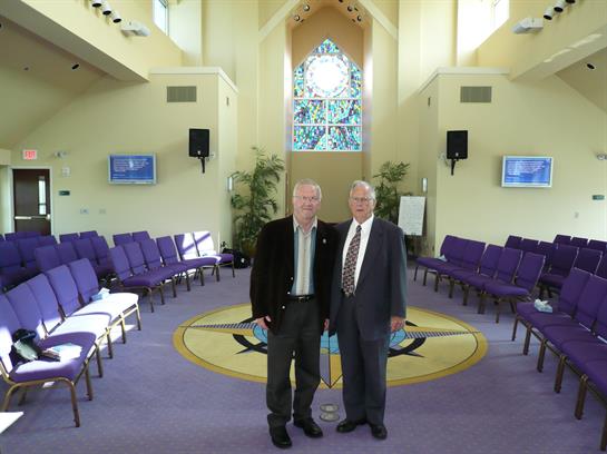Two men pose for a picture inside a brightly lit church with vibrant stained glass.