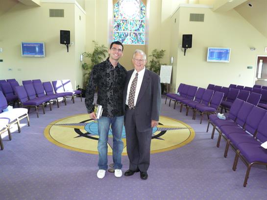 Two men stand smiling in a well-lit hall featuring purple seating and an intricate window design.