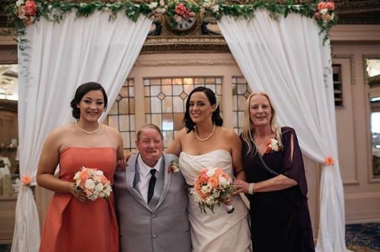Four women in formal attire pose for a joyful photo at a wedding, smiling and holding bouquets.