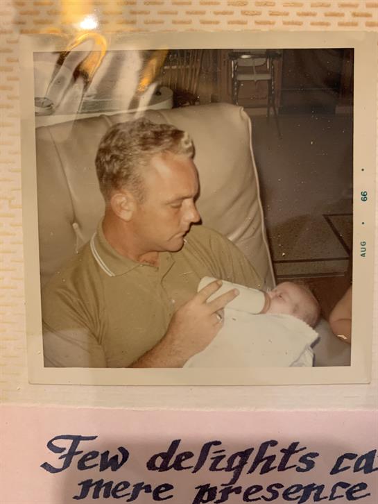 A father gently holds and feeds his tiny baby in a warm, inviting living room filled with nostalgia.