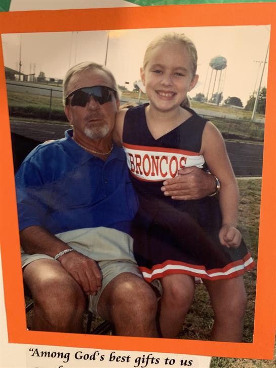 A young girl in a cheerleading outfit sits happily on a man's lap. They enjoy a sunny day.