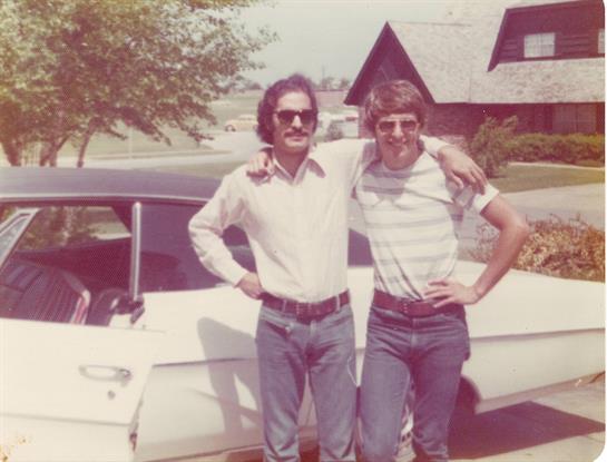 Two men stand proudly with their arms around each other next to a vintage car on a sunny day.