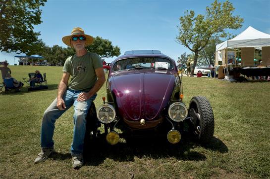 A man in a wide-brimmed hat sits by a vintage purple car under a clear sky.