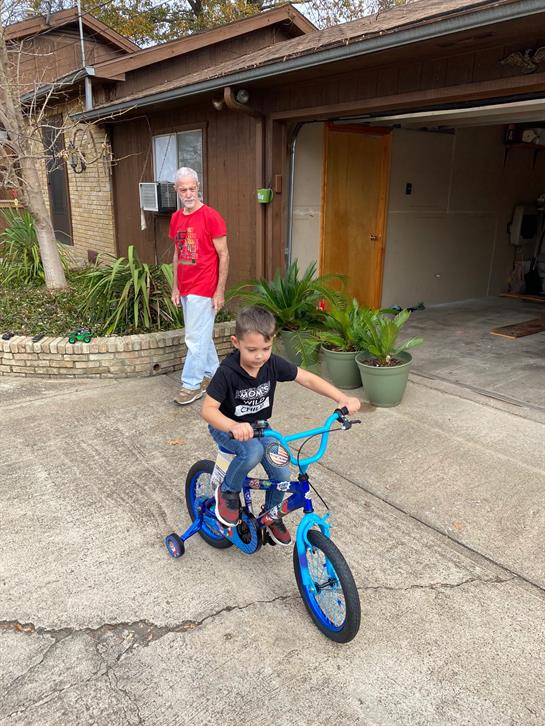 A boy skillfully rides a blue bicycle in a driveway under the watchful eye of an adult.