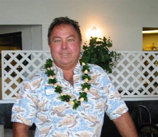 Man stands proudly with a bright smile, wearing a floral shirt and a lei at a celebratory event.