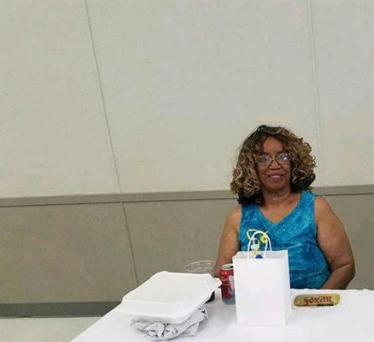 An elderly woman in a bright blue dress sits at a community center table enjoying snacks.