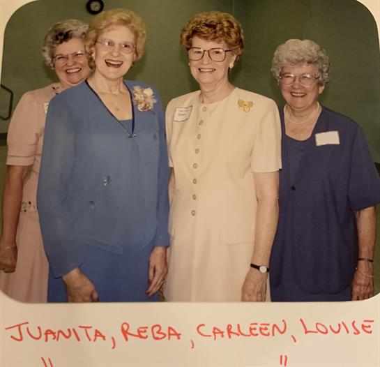 Four women stand together joyfully at a gathering, each wearing colorful outfits and sharing smiles.
