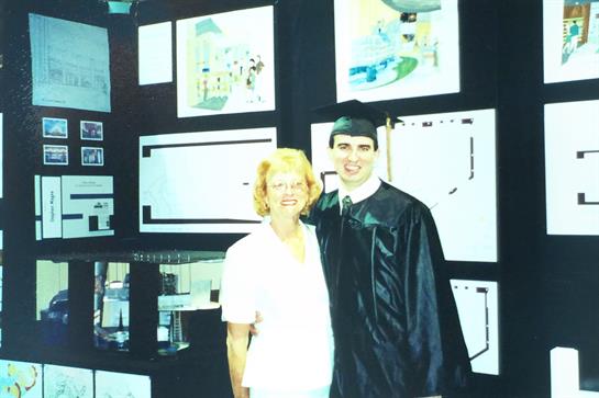 A proud graduate stands beside a family member in front of a wall displaying creative projects.