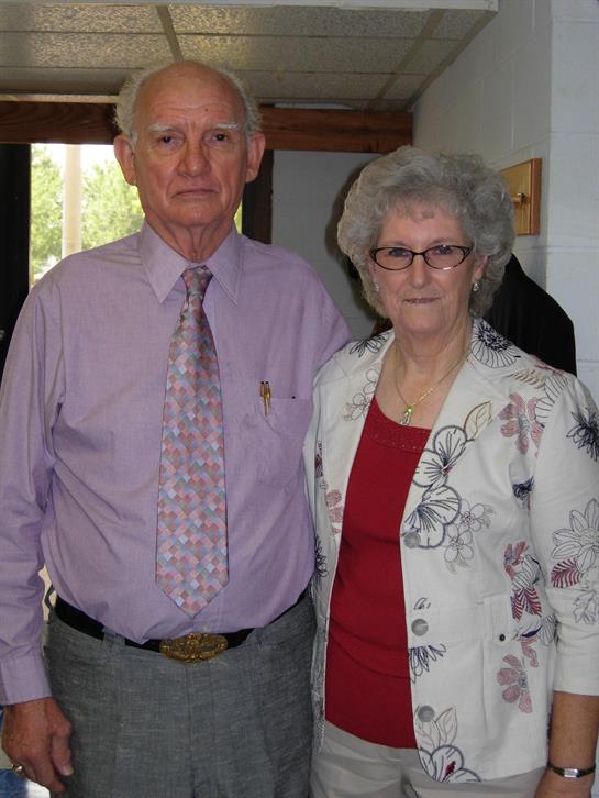 An elderly couple poses together, dressed in formal attire, at a family gathering indoors.