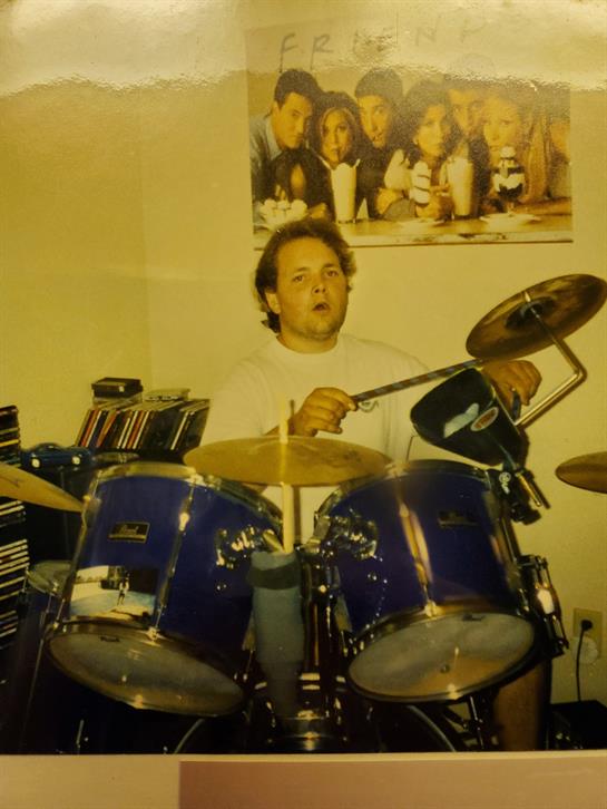 Musician plays drums energetically in a cozy home studio filled with instruments and friends.