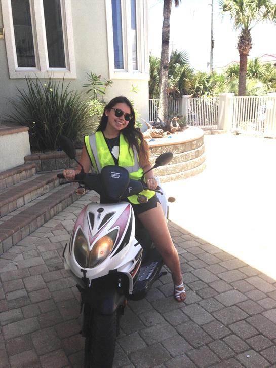 Woman wearing a reflective vest enjoys a sunny day while sitting on her scooter in front of a house.