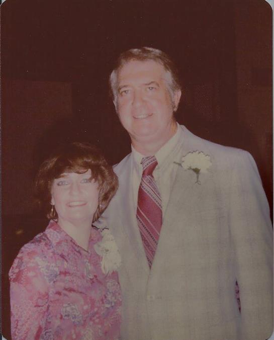 A man and woman pose together cheerfully during a joyful event, dressed formally with flowers.