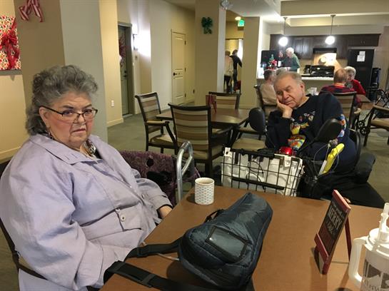 Two seniors sit at a table in a community center, engaged in conversation and reminiscing.