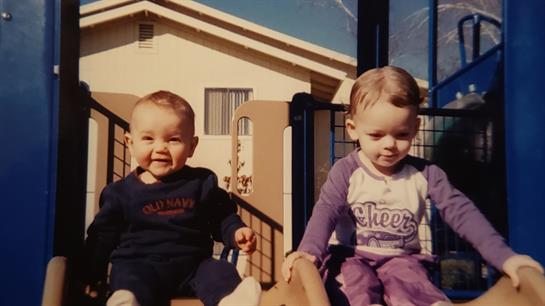 Two young children play on a slide together, smiling and enjoying a sunny day outdoors.