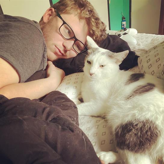 A person with glasses lies comfortably next to a white cat on a bed, enjoying a quiet moment.