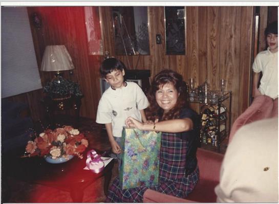 A woman smiles while holding a wrapped gift as a child stands nearby during a cozy indoor gathering.